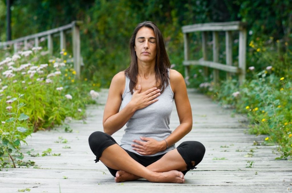 Mujer meditando al aire libre con las manos en el pecho y abdomen, practicando respiración consciente.