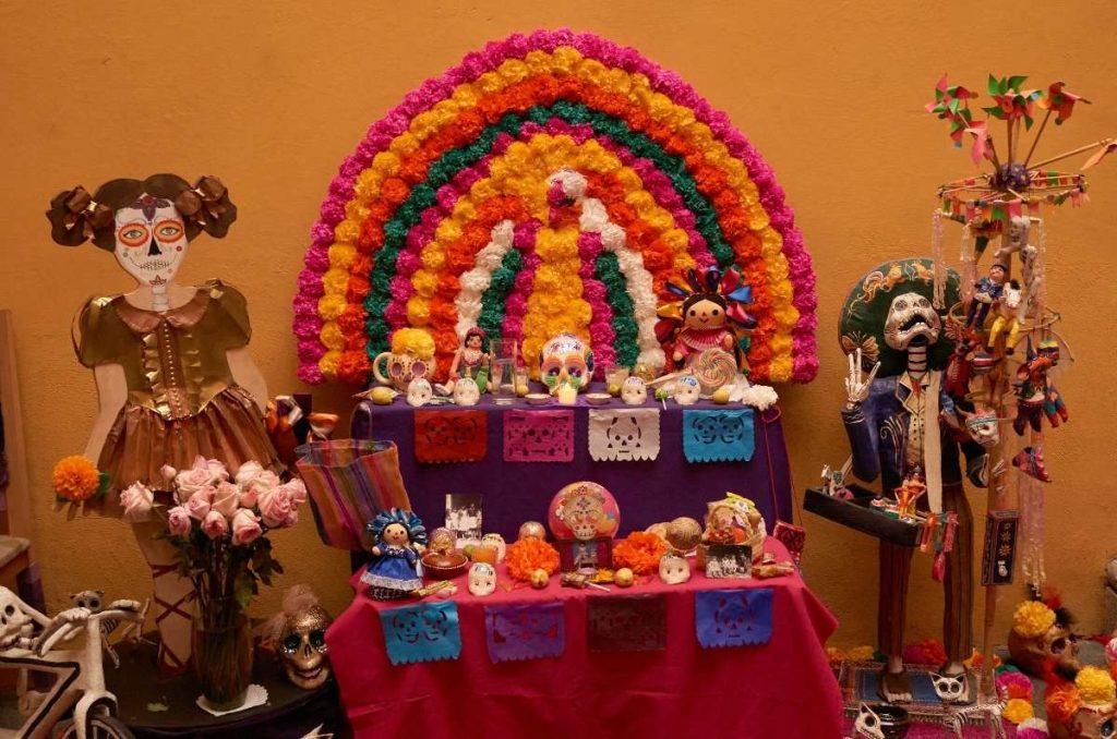 Vista de altares y ofrendas de Día de Muertos con flores, velas y calaveritas decorativas.
