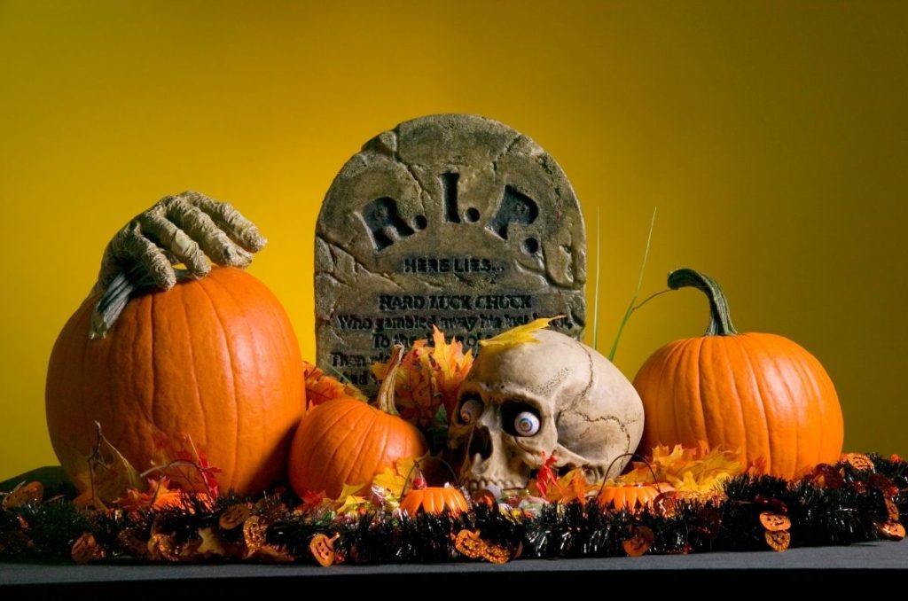 Lápida decorativa con tierra, cráneos, calabazas y flores naranjas para decorar casa de terror con temática de Halloween