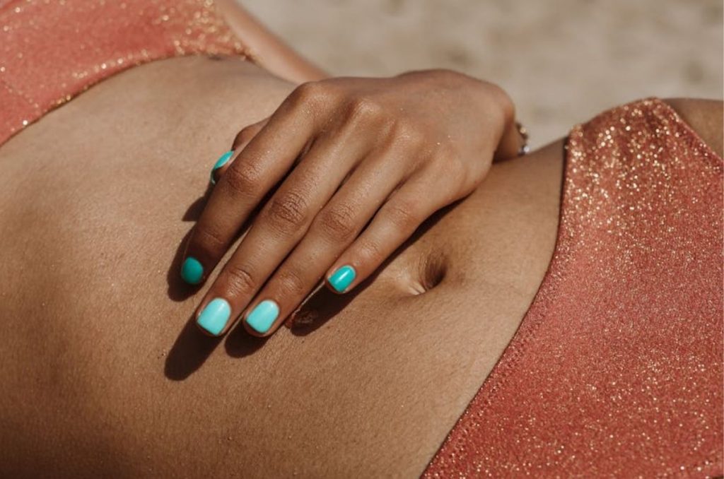 Abdomen bronceado recostado en la playa con ropa interior naranja; una mano reposa sobre el vientre mostrando uñas color turquesa.