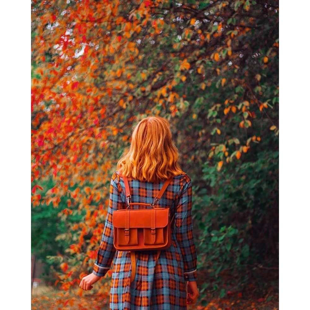 Chica en bosque otoñal con abrigo de tartán y mochila de piel a la espalda con cabello suelto corto cobrizo