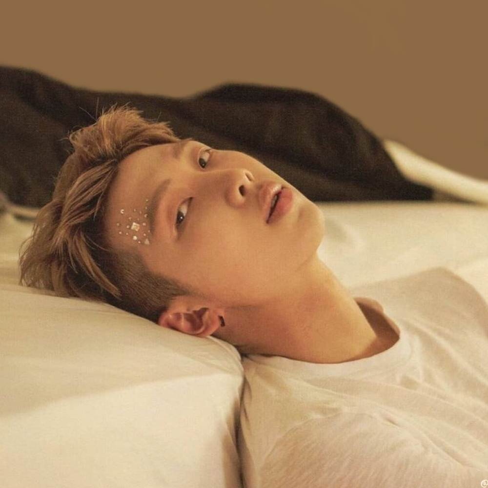 RM sentado en el suelo y recostando su cabezo sobre una cama blanca