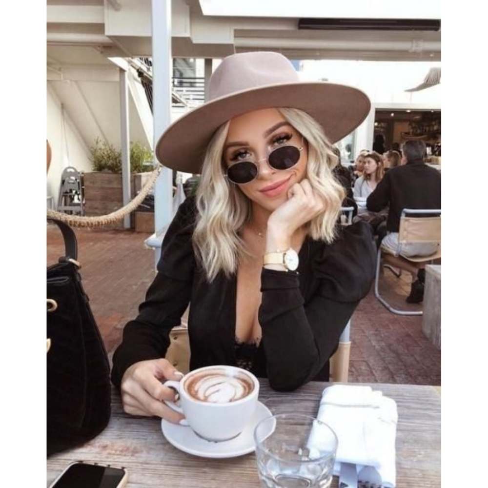 Chica con sombrero de ala ancha en color tierra y lentes oscuros tomando café en un día de otoño con outfit negro