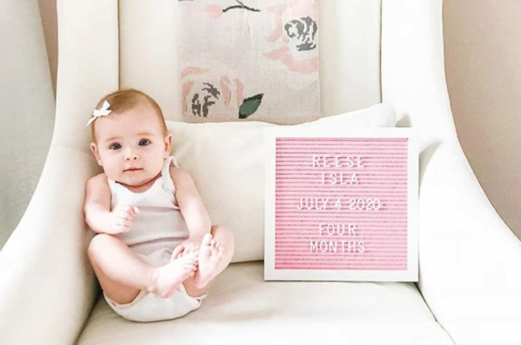 Bebé sentada en un sillón blanco con cojines de flores rosas, junto a un pizarrón rosa con letras blancas que muestran su nombre, fecha de nacimiento y los meses que cumple. Idea original para la celebración mensual de tu bebé
