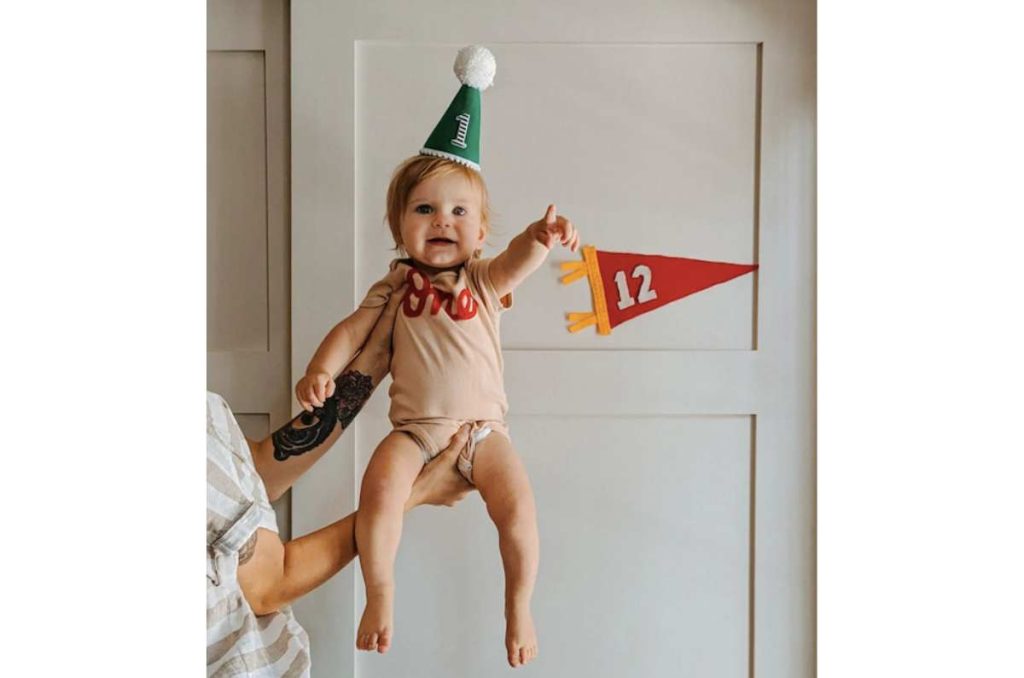 Bebé de un año con mameluco beige que dice “One” en letras rojas y gorro de fiesta verde con el número 1, siendo cargado por alguien frente a un fondo blanco con banderín rojo del número 12. Idea de fotos para la celebración mensual de tu bebé.