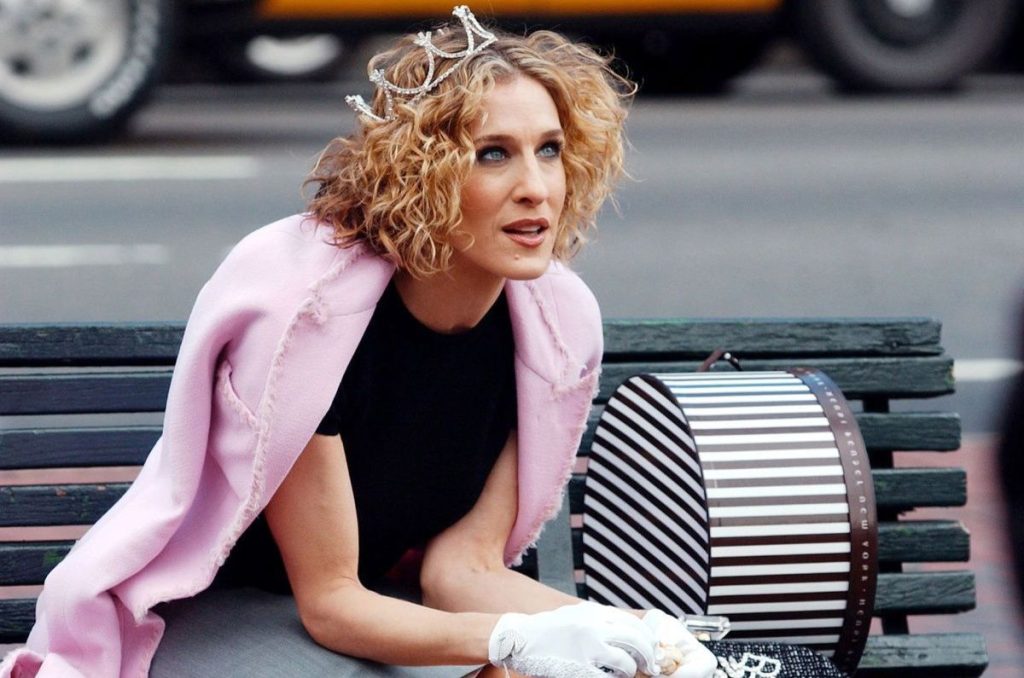 Carrie Bradshaw sentada en una banca de la ciudad, usando un outfit negro con un saco rosa pastel, observando algo a lo lejos.