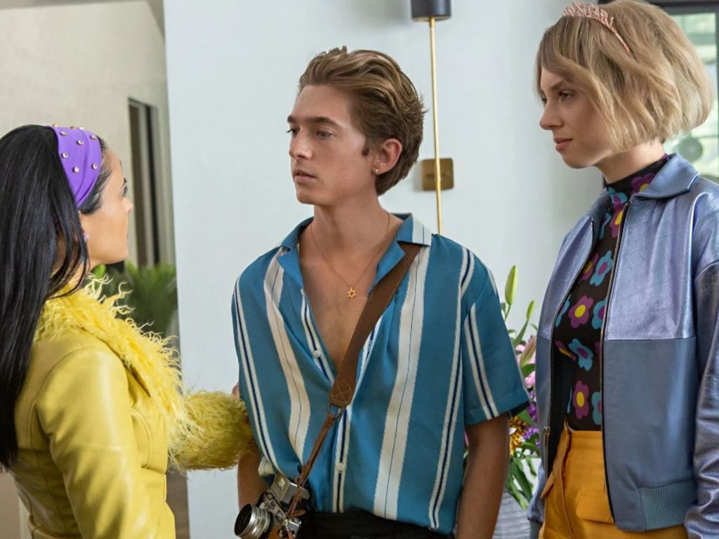 Una chica de cabello negro con diadema morada vestida de amarillo habla con un chico rubio vestido con camisa azul con blanco y pantalones negros, mientras otra chica rubia vestida con blusa negra con flores de colores, chamarra azul y pantalones amarillos los observa
