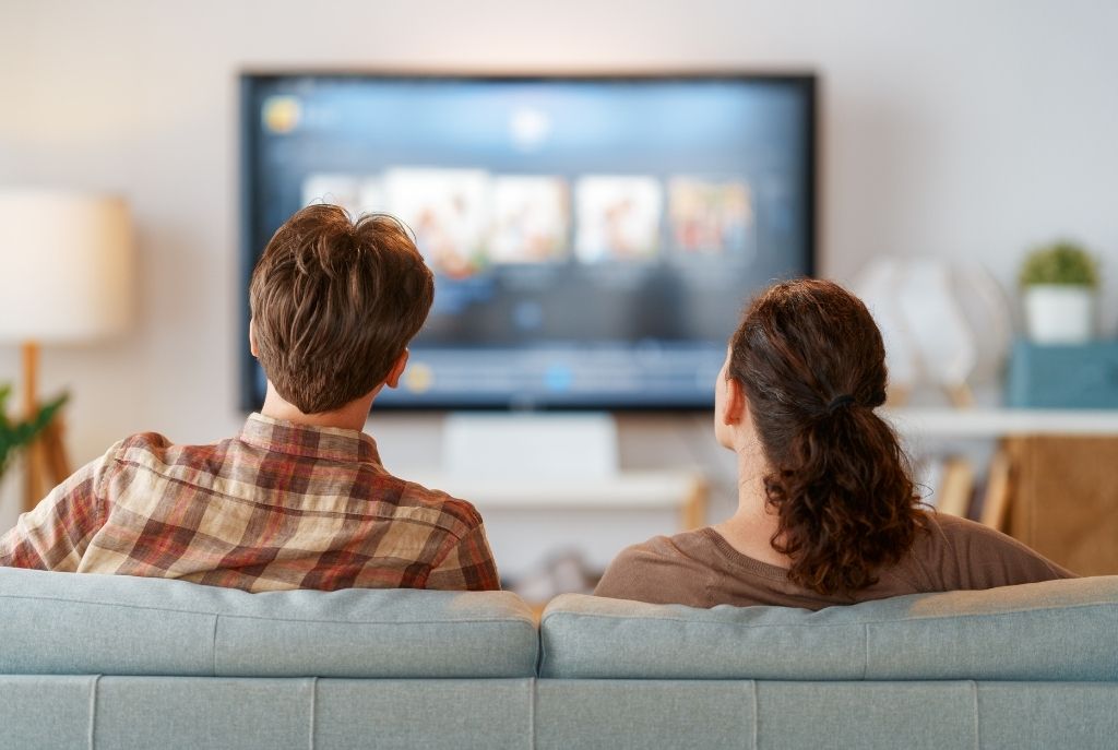 Pareja sentada viendo televisión juntos en habitación de motel, compartiendo un momento cercano y divertido