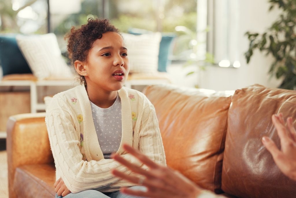 Una niña sentada en un sillón con expresión de confusión mientras un adulto le explica de forma tierna y simple cómo llegan los bebés al mundo