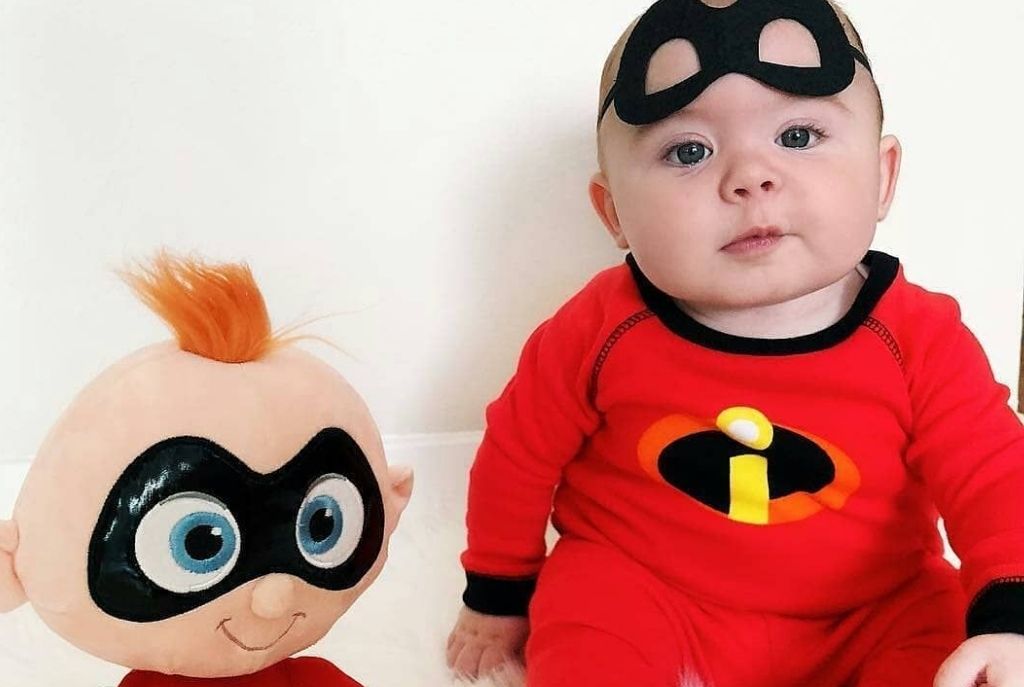 Bebé sentado con disfraz de Jack-Jack de Los Increíbles, acompañado a su lado izquierdo por un peluche del mismo personaje. Ideal para Halloween para tu bebé con un look de superhéroe adorable