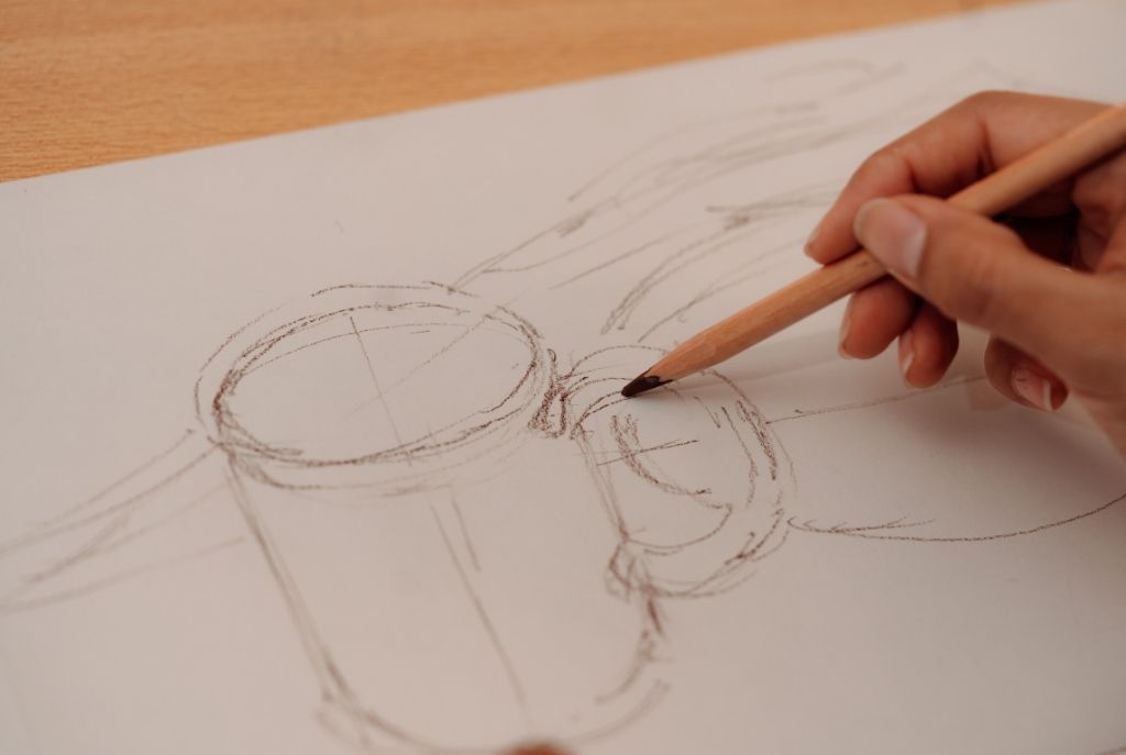 Una mano que sostiene un lápiz y dibuja una taza en una hoja blanca, disfrutando de un juego casero de pictionary donde lo divertido es intentarlo, no ser un artista
