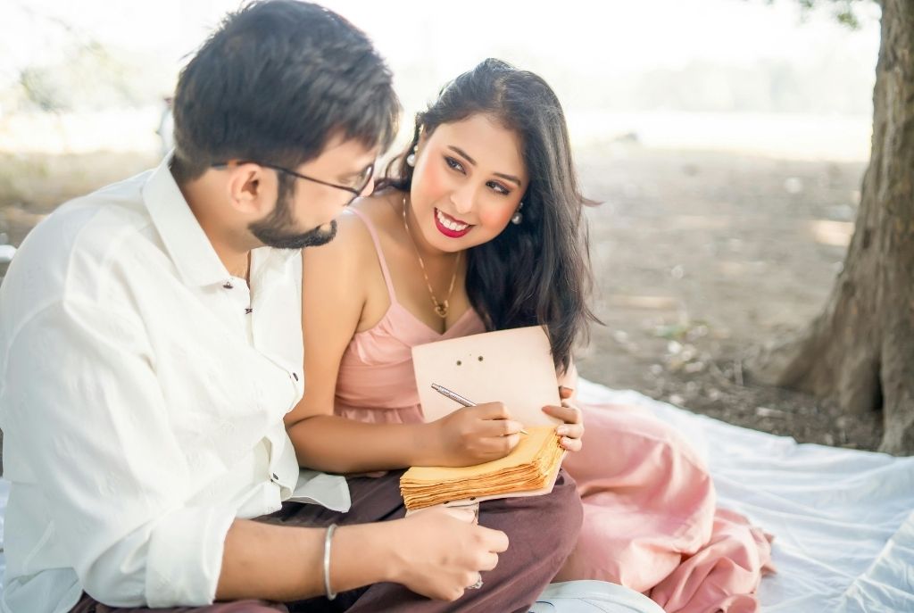 Pareja sentada en una sábana en el parque, ella con vestido rosa y una libreta en mano lista para escribir mientras él la mira atento y sonriente, disfrutando de juegos en pareja de manera sencilla y divertida