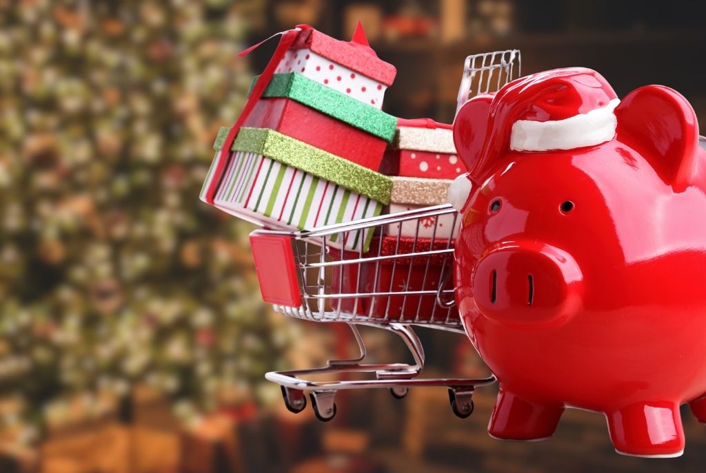 Cochinito rojo de ahorro con un gorro de santa, junto a un carrito de compras con regalos navideños frente a un árbol de Navidad con luces 