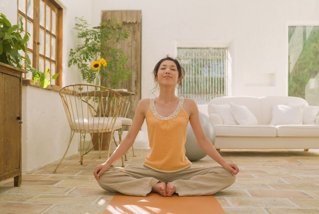 Mujer haciendo yoga en casa, sentada en un tapete naranja con piernas abiertas tipo mariposa, ojos cerrados, vistiendo blusa de tirantes naranja y pants beige