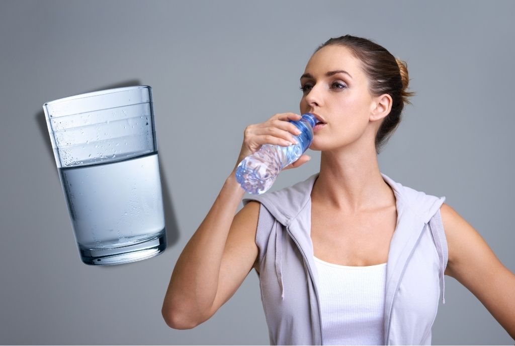 Mujer con chongo, blusa blanca y chaleco deportivo tomando agua de una botella para desinflamar el vientre, junto a un vaso grande de agua sobre un fondo sólido