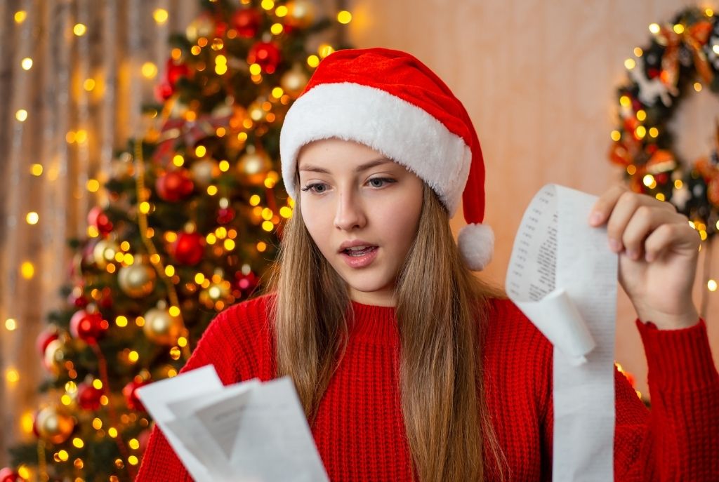 Mujer rubia de ojos azules con suéter rojo y gorro de Santa Claus mirando sorprendida unos tickets de compras frente a un árbol de Navidad iluminado