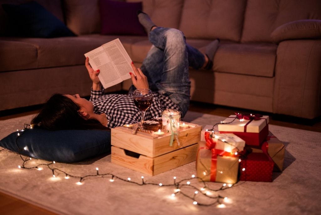 Chica acostada en la alfombra de su sala leyendo un libro, rodeada de luces LED y regalos, en un ambiente navideño acogedor