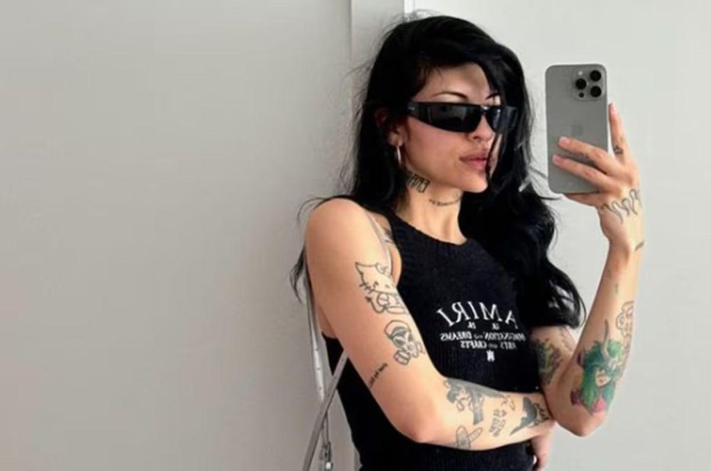 Cazzu tomándose una foto en el espejo con su celular, usando lentes oscuros y ropa negra