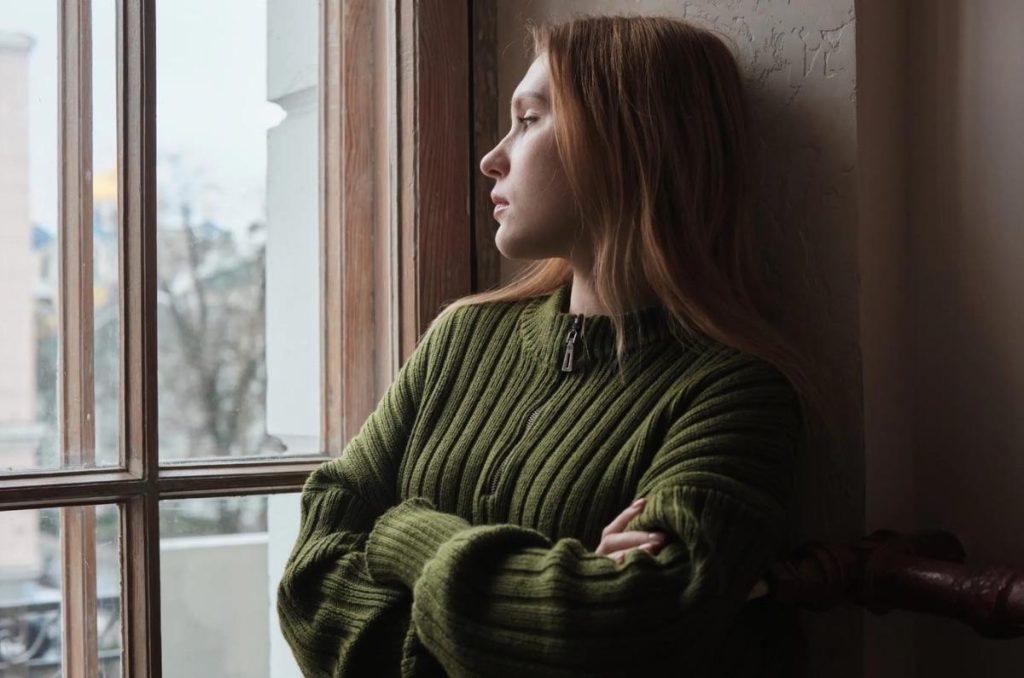 Mujer recargada en la pared con los brazos cruzados, mirando hacia una ventana con expresión pensativa, tiene miedo al matrimonio