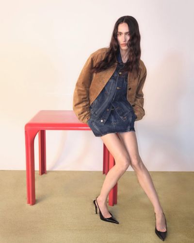 Modelo sentada junto a una mesa roja, con mini vestido de mezclilla, chamarra café y tacones negros, sobre fondo claro.