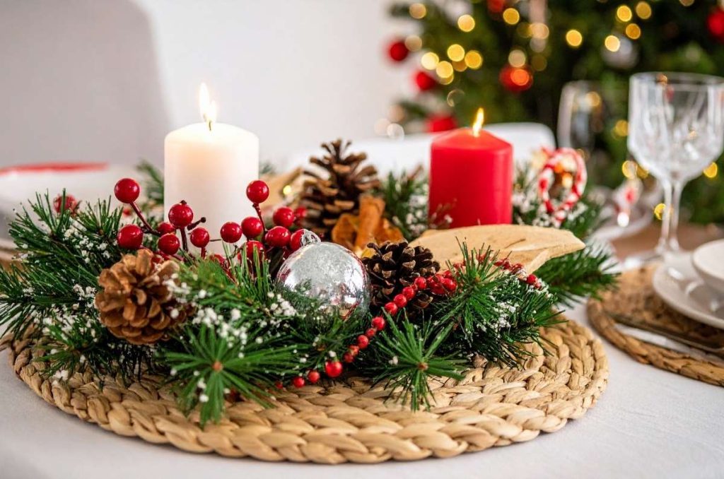 Centro de mesa navideño con velas roja y blanca, piñas, ramas de pino y esferas plateadas sobre base de mimbre, con luces del árbol de Navidad desenfocadas al fondo.