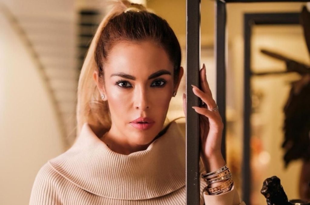 Cristina Beyruti con vestido beige de cuello alto y cabello en coleta, recargada en una estructura metálica dentro de un interior elegante con iluminación cálida.