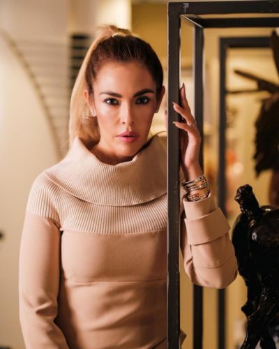 Cristina Beyruti con vestido beige de cuello alto y cabello en coleta, recargada en una estructura metálica dentro de un interior elegante con iluminación cálida.