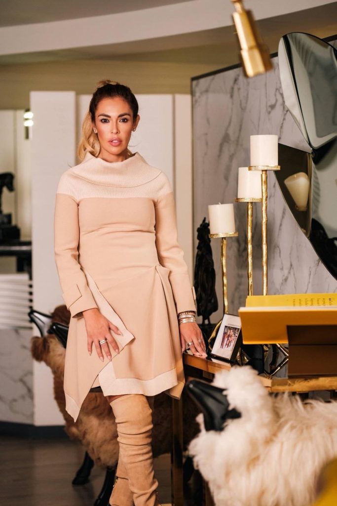 Cristina Beyruti de cuerpo completo con vestido beige y botas altas, de pie junto a una consola con velas decorativas y objetos dorados en una sala de estilo moderno.