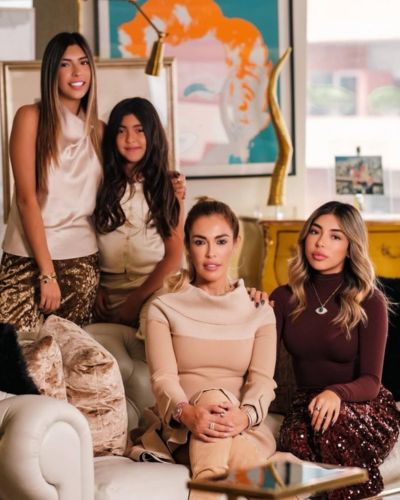 Cristina Beyruti posa con sus hijas en una sala con sofá blanco y decoración sofisticada; una está sentada al centro y las otras tres están de pie y sentadas a su alrededor.
