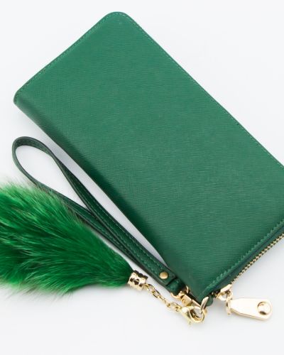 Cartera larga verde sobre fondo blanco, con cierre y correa; a un lado cuelga una borla de pelo verde con herrajes dorados.