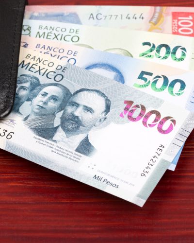 Monedas y billetes de pesos mexicanos extendidos frente a una cartera gris, sobre fondo neutro.