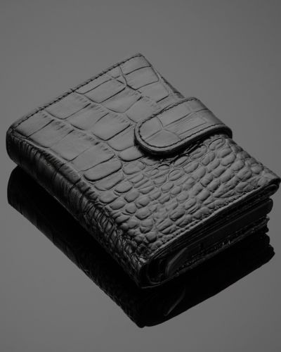 Cartera pequeña negra con textura tipo piel grabada y broche, colocada sobre una superficie oscura con reflejo.