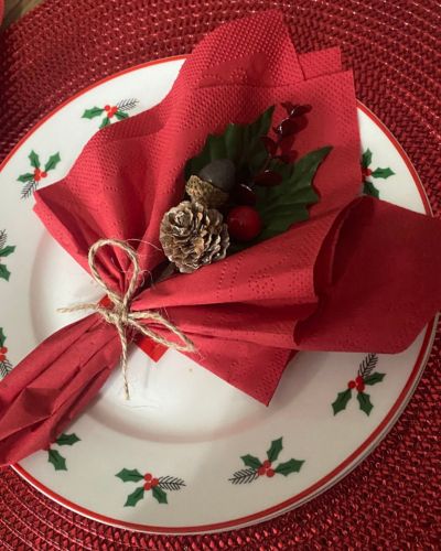 Servilleta roja doblada y amarrada con yute sobre un plato con motivos navideños; encima lleva un adorno con piña de pino, hojas verdes y pequeños detalles decorativos.