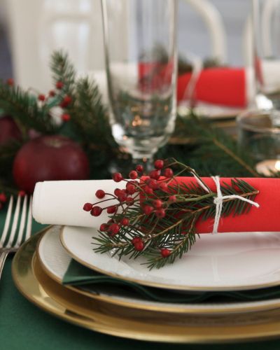 Servilleta enrollada en rojo y blanco sobre plato, adornada con ramitas verdes y frutos rojos amarrados con hilo; alrededor hay vajilla navideña y copas.