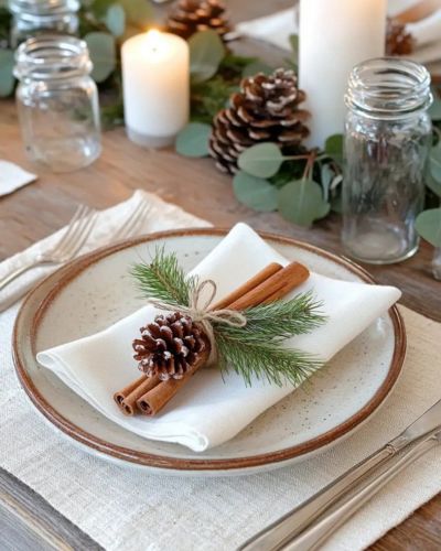 Servilleta blanca doblada sobre plato beige, decorada con una ramita de pino, una piña de pino y dos rajas de canela sujetas con cordel; al fondo hay velas y frascos de vidrio.