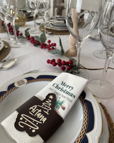 Mesa formal con copas y platos; servilleta blanca con texto “Merry Christmas and happy New Year” colocada en un servilletero oscuro con letras “All in a while”, con ramas y frutos rojos decorando el centro.