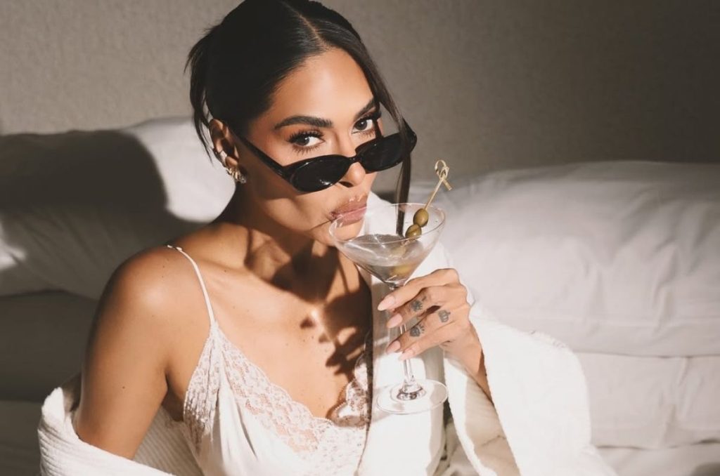 Galilea Montijo recostada sobre una cama con lencería clara y una bata blanca; lleva lentes de sol, sostiene una copa tipo martini y frente a ella hay un plato con botanas sobre la cama.