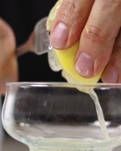 Primer plano de una mano exprimiendo un limón con un tenedor, dejando caer el jugo en un recipiente de vidrio transparente.