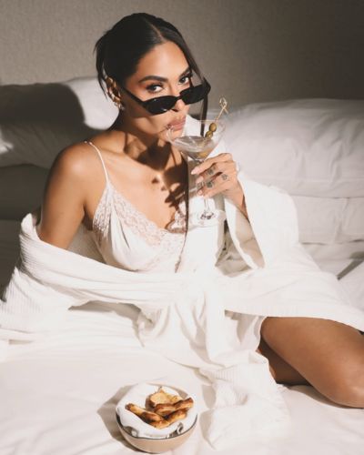 Galilea Montijo  recostada sobre una cama con lencería clara y una bata blanca; lleva lentes de sol, sostiene una copa tipo martini y frente a ella hay un plato con botanas sobre la cama.