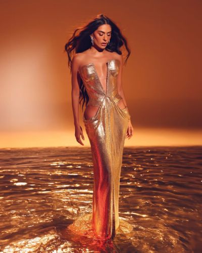 Galilea Montijo de pie en agua poco profunda con un vestido largo dorado metalizado de escote pronunciado; el fondo y la luz son cálidos en tonos naranja, y su cabello largo ondea mientras mira hacia abajo.