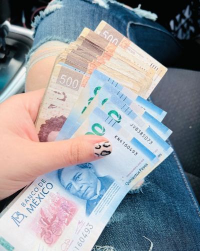 Persona sentada en un auto sostiene varios billetes mexicanos de 500, 200 y otras denominaciones; se ve mezclilla rasgada en la pierna y uñas con diseño.
