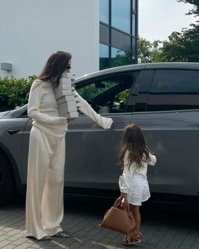Foto en plano general de una mujer con iun conjunto beige de pantalón y sudadera abriendo la puerta de una camioneta gris y a su lado una niña con un conjunto de blusa y shot en color blanco
