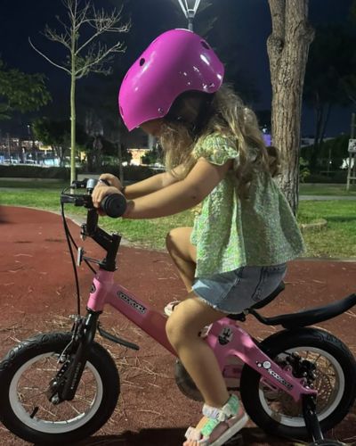 Niña con casco rosa montando una bicicleta infantil rosa en un parque por la noche.