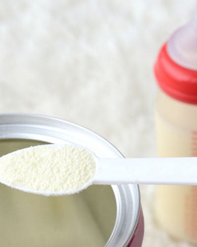 Cucharita con leche en polvo sobre una lata abierta; al fondo se aprecia un biberón desenfocado.