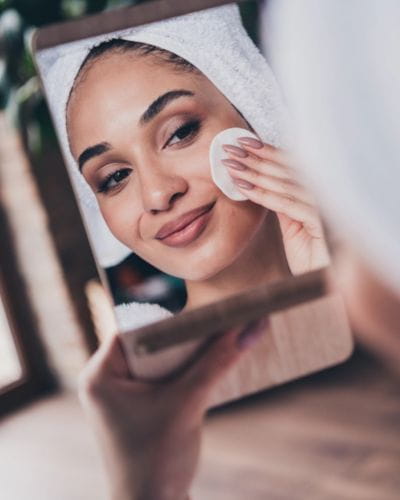 Mujer con toalla en la cabeza limpiando su rostro con un algodón frente al espejo.