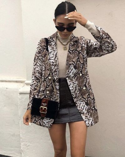 Foto de mujer de pie con outfit semi formal: mini falda negra, blazer estampado de serpiente, blusa de cuello alto color beige y bolso negro