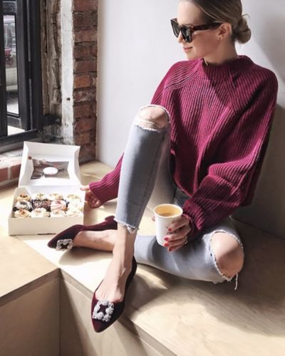Mujer sentada junto a una ventana con café; usa bailarinas negras con pedrería y jeans rotos.