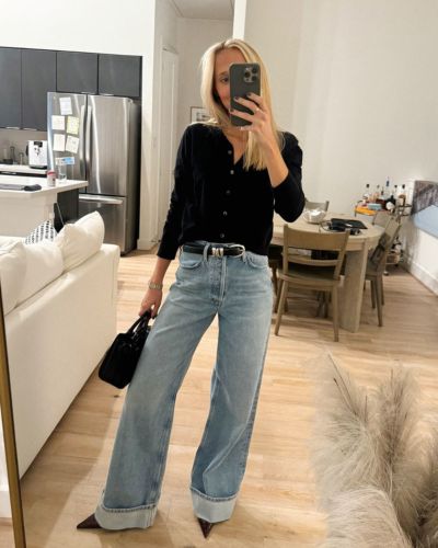 Mujer frente al espejo con blusa negra y jeans wide leg; lleva tacones negros de punta.