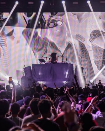 DJ en escenario del Tecate Emblema con público levantando las manos y luces blancas iluminando el concierto.