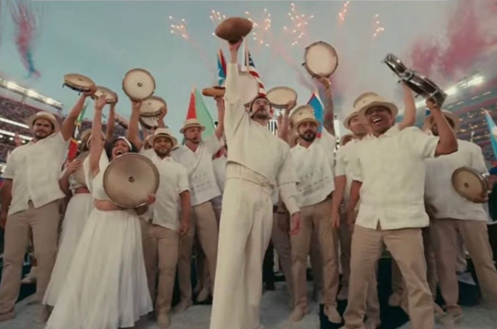 Bad Bunny durante el medio tiempo del Super Bowl con vestuario en tono Pantone crema, acompañado de músicos con panderos y fuegos artificiales al fondo.